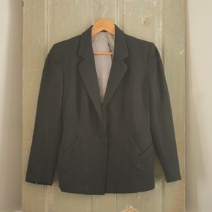 May include: A black blazer with a single button closure and two decorative pockets on the front. The blazer is hanging on a wooden hanger against a light green wooden door.