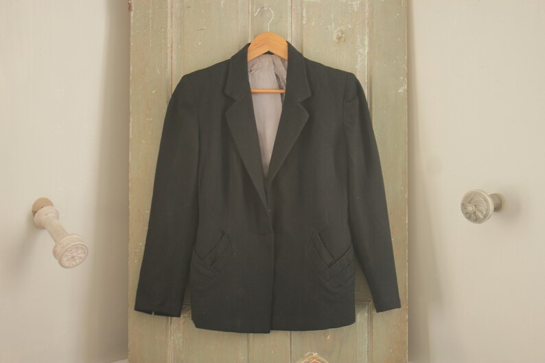 May include: A black blazer with a single button closure and two decorative pockets on the front. The blazer is hanging on a wooden hanger against a light green wooden door.