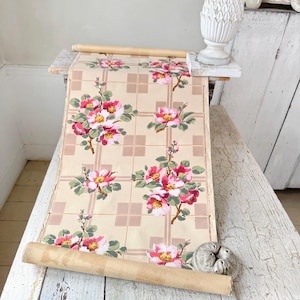 May include: A roll of vintage wallpaper featuring a repeating floral pattern of pink and white roses with green leaves on a beige background with a grid design. The wallpaper is rolled around a wooden dowel.