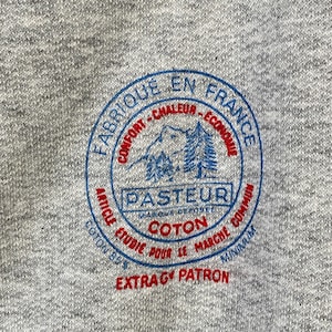 Pode incluir: Uma camisola cinzenta com um logotipo circular azul e vermelho que diz "Fabrique en France" e "Pasteur" com uma montanha e &aacute;rvores dentro do c&iacute;rculo. O logotipo tamb&eacute;m diz "Confort - Chaleur - Economique" e "Article Etudie Pour Le Marche Commun" e "Coton" e "Extra G Patron".