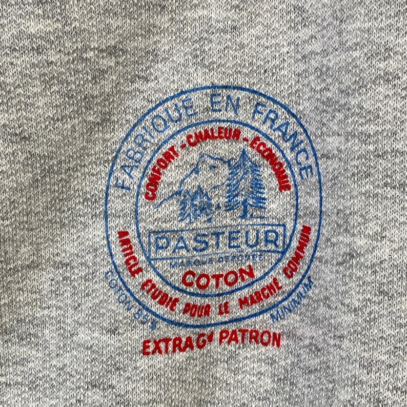 Pode incluir: Uma camisola cinzenta com um logotipo circular azul e vermelho que diz "Fabrique en France" e "Pasteur" com uma montanha e &aacute;rvores dentro do c&iacute;rculo. O logotipo tamb&eacute;m diz "Confort - Chaleur - Economique" e "Article Etudie Pour Le Marche Commun" e "Coton" e "Extra G Patron".