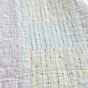 May include: Close-up of a patchwork quilt with a variety of patterns. Sections include white fabric with vertical yellow stripes and colorful dots, and sections with pink floral patterns. The quilt is made of soft, textured fabric.