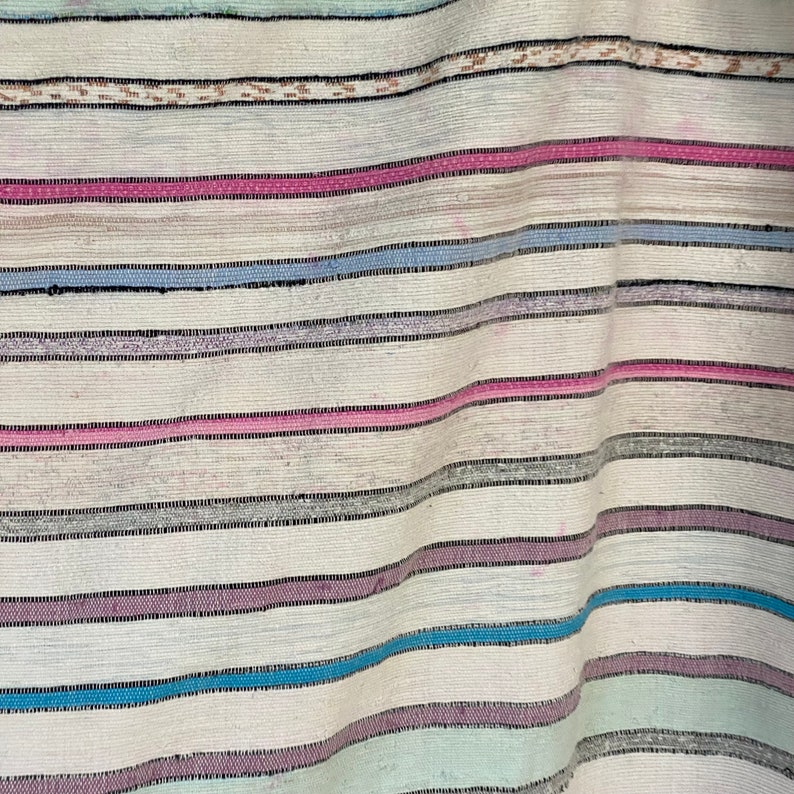 May include: A white woven fabric with horizontal stripes in shades of pink, blue, and gray. The fabric is folded and has a textured surface.