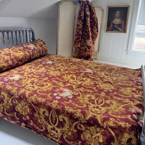Könnte beinhalten: Ein Bett mit einer bordeauxroten und goldfarbenen Bettdecke und einem Zierkissen mit floralem Muster. Das Bett hat einen dunklen Metallrahmen. Ein passender Stoff mit floralem Muster hängt an einem Paravent hinter dem Bett.