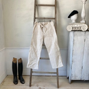 May include: A pair of white linen pants with thin brown stripes. The pants have a tie at the waist and are hanging on a wooden ladder.