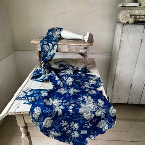 May include: A blue and white floral fabric with a white background and blue flowers. The fabric is draped over a white table with a white vase on top.