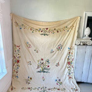 May include: A large, cream-colored tablecloth with intricate floral embroidery in shades of pink, blue, green, and red. The tablecloth has a delicate white fringe border.