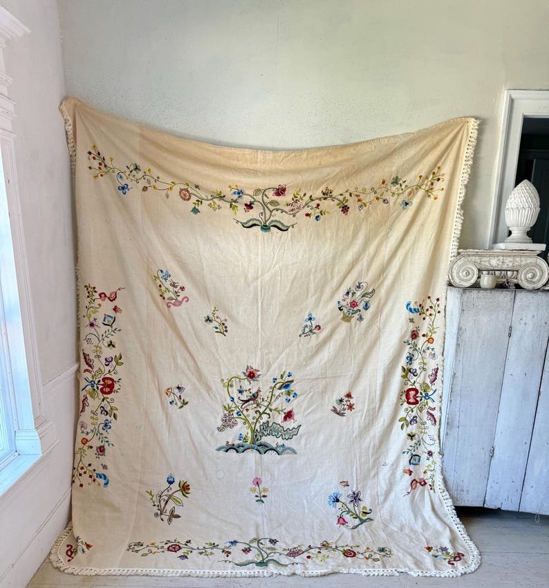 May include: A large, cream-colored tablecloth with intricate floral embroidery in shades of pink, blue, green, and red. The tablecloth has a delicate white fringe border.