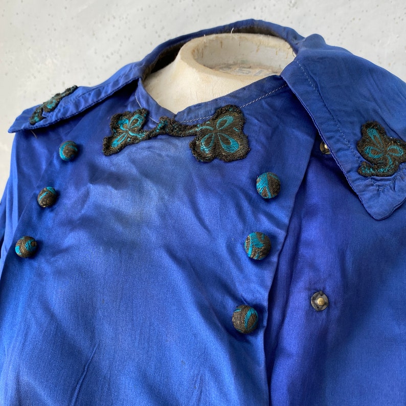 May include: Close-up of a vintage cobalt blue blouse. The blouse features a unique collar and decorative black and teal embroidered accents. Round buttons with teal and black detailing adorn the front. The fabric has a subtle sheen.