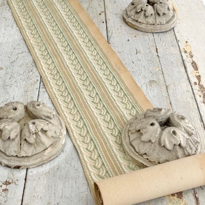 Pode incluir: Um rolo de papel de parede vintage com um padrão repetitivo de listras verticais em bege, branco e verde claro. Também estão presentes três ornamentos circulares de pedra.