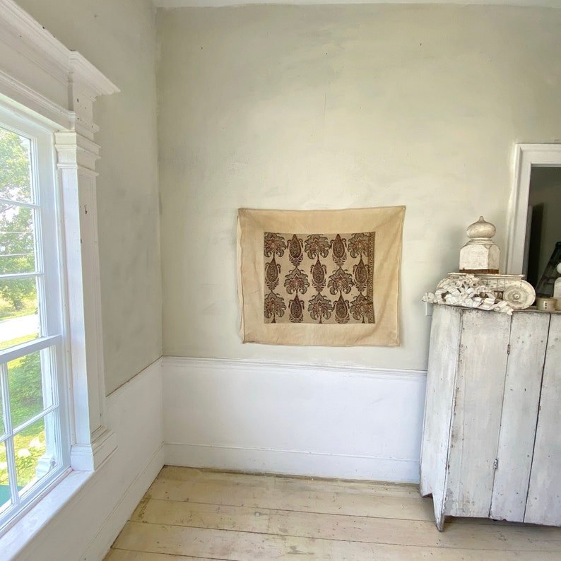Pu&ograve; includere: Un muro beige con una cornice bianca e un armadio in legno bianco. Un arazzo color crema con un motivo floreale marrone &egrave; appeso al muro.