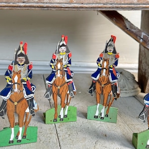 May include: Wooden toy soldiers on horseback. Each soldier is dressed in a blue and red uniform with a tall hat. The horses are brown with white legs, and the soldiers are mounted on green bases. The toys are arranged on a weathered wooden surface.