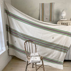 May include: A large white linen tablecloth with colorful stripes and the initials "EC" embroidered in the corner. The tablecloth is draped over a wooden chair and a small table.