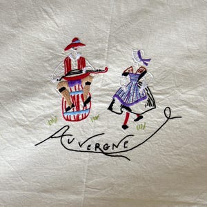 May include: Embroidered linen tablecloth featuring a scene of two figures in traditional dress, one playing a musical instrument. The scene is embroidered in red, blue, green, and black thread. The text "AUVERGNE" is embroidered in black thread below the figures.