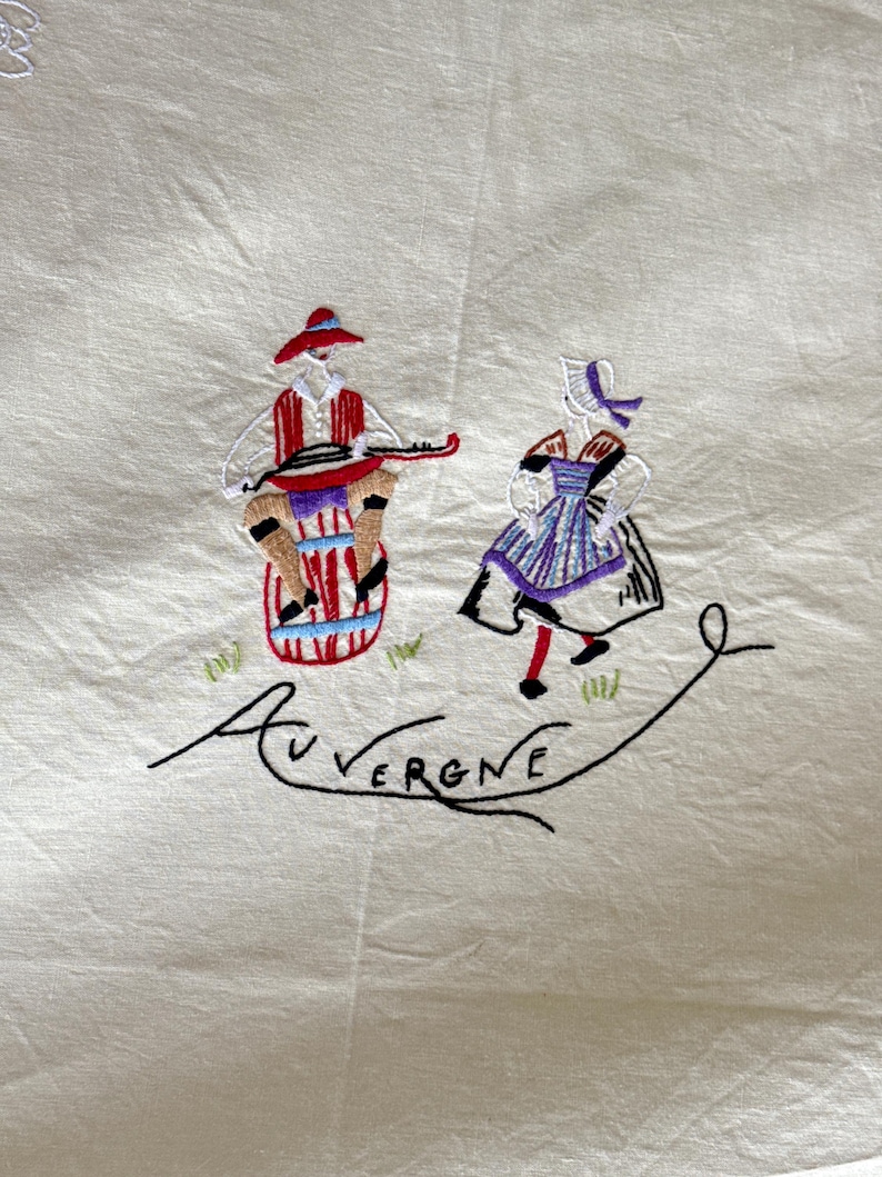 May include: Embroidered linen tablecloth featuring a scene of two figures in traditional dress, one playing a musical instrument. The scene is embroidered in red, blue, green, and black thread. The text "AUVERGNE" is embroidered in black thread below the figures.