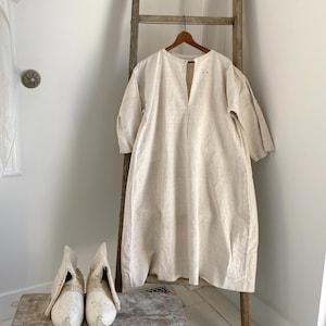 Puede incluir: Un vestido de lino color crema con cuello en V y mangas cortas cuelga de una percha de madera. Dos hormas de zapatos de madera se asientan sobre un pequeño taburete de madera frente a una escalera de madera. El vestido tiene un diseño sencillo y relajado.