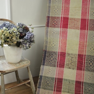 Puede incluir: Una alfombra tejida vintage con un patrón a cuadros en tonos rojos, azules y crema. La alfombra está colgada en una pared en una habitación con una silla de madera y un jarrón de flores secas.