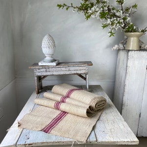 Puede incluir: Un banco de madera rústico con un acabado blanqueado se encuentra sobre una mesa blanca con un camino de mesa de tela a rayas beige y rojo. El banco está decorado con una piña de cerámica blanca.