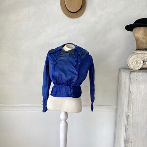 May include: A vintage cobalt blue jacket with long sleeves and a fitted waist, displayed on a white dress form. The jacket features decorative buttons and embroidered detailing on the collar. A straw hat hangs in the background.