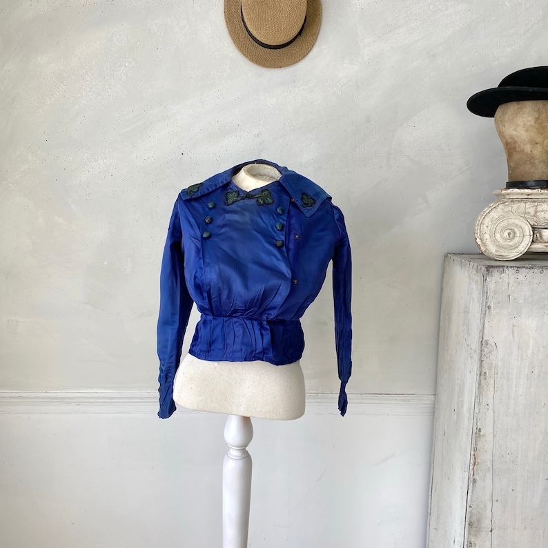 May include: A vintage cobalt blue jacket with long sleeves and a fitted waist, displayed on a white dress form. The jacket features decorative buttons and embroidered detailing on the collar. A straw hat hangs in the background.