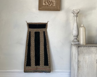 May include: A black velvet skirt with gold embroidery and a red, white, and blue trim hangs on a white wall. A wooden frame with a carved design hangs above the skirt. A white painted pedestal with a bottle on top sits on a white painted cabinet.