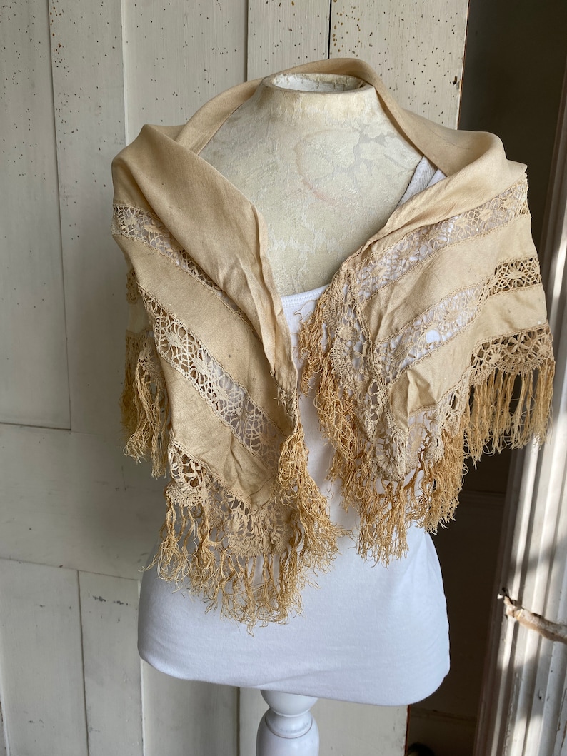 May include: A cream-colored lace shawl with fringe trim. The shawl is displayed on a white mannequin.