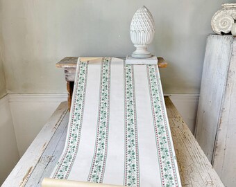 May include: A roll of vintage wallpaper with a cream background and vertical stripes. The stripes are bordered by green and red floral patterns. The wallpaper is displayed on a distressed white table.