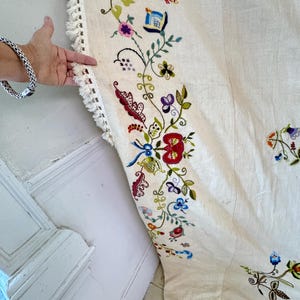 May include: A white linen tablecloth with a hand-stitched embroidered border featuring colorful floral designs. The embroidery includes flowers, leaves, and vines in shades of red, blue, green, pink, and yellow.