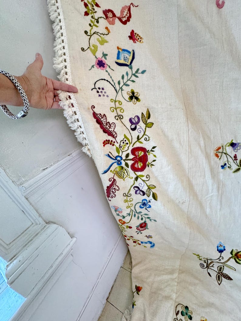 May include: A white linen tablecloth with a hand-stitched embroidered border featuring colorful floral designs. The embroidery includes flowers, leaves, and vines in shades of red, blue, green, pink, and yellow.