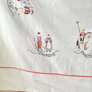 May include: White linen tablecloth with red embroidered stitching around the edges. The tablecloth features colorful embroidered figures representing different regions of France, including "AINE", "LOURAINE", and "PROVEN.