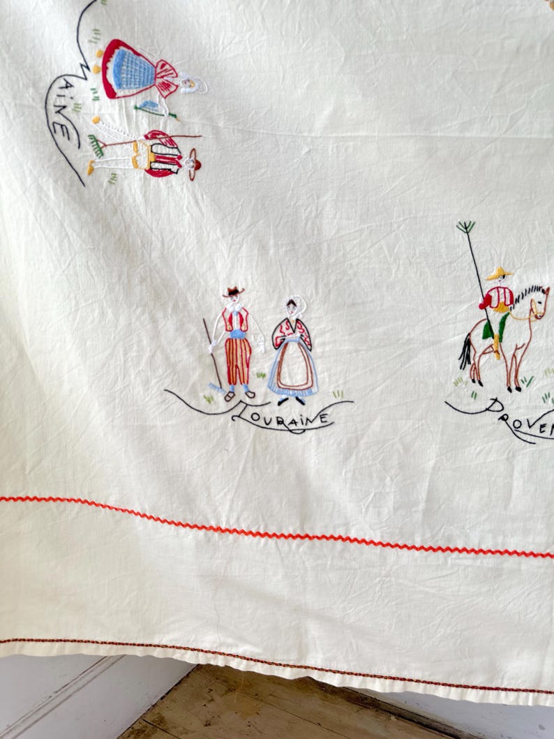 May include: White linen tablecloth with red embroidered stitching around the edges. The tablecloth features colorful embroidered figures representing different regions of France, including "AINE", "LOURAINE", and "PROVEN.