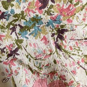 May include: Close-up of a white fabric with a floral pattern. The pattern features pink, blue, and green flowers on a white background.
