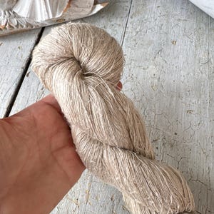 Puede incluir: Un ovillo de hilo retorcido de color natural, sostenido en una mano. El hilo es de color beige claro y parece estar hecho de una fibra natural. El fondo incluye una superficie de madera clara desgastada y un elemento decorativo blanco.