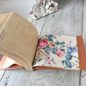 May include: An open sample book with a tan cover featuring a page of floral wallpaper. The wallpaper displays pink, yellow, and blue flowers with green leaves on a cream background. The adjacent page contains printed text and the code 7186 C.