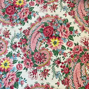 May include: Close-up of a floral fabric with a paisley design. The fabric features a white background with a repeating pattern of pink roses, yellow and blue flowers, and green leaves. The paisley elements are in pink and red.
