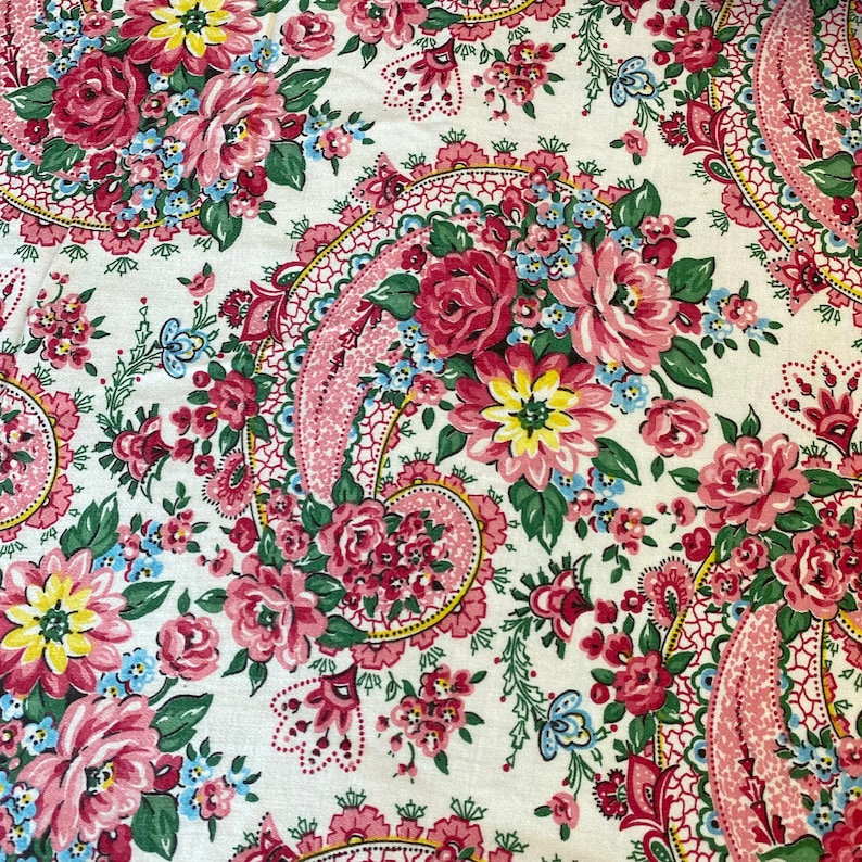 May include: Close-up of a floral fabric with a paisley design. The fabric features a white background with a repeating pattern of pink roses, yellow and blue flowers, and green leaves. The paisley elements are in pink and red.