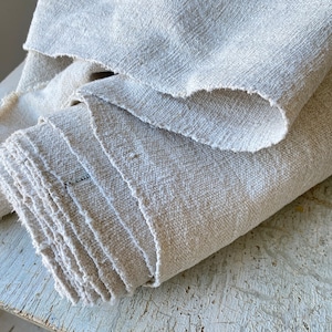 May include: A close-up of a stack of natural linen fabric. The fabric is a light beige color and has a rough, textured surface. The fabric is folded in a neat pile.
