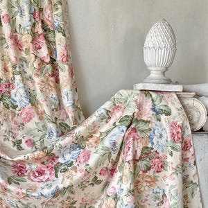 Könnte beinhalten: Cremefarbener Blumendruckstoff mit großen rosa, blauen und pfirsichfarbenen Rosen und grünen Blättern. Der Stoff ist drapiert, mit einem weißen, geschnitzten, ananasförmigen Dekorationsgegenstand im Hintergrund.