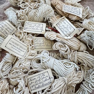 May include: A pile of natural fiber twine, with various sizes and lengths, labeled with handwritten tags in French. The tags indicate the type of twine, the quantity, and the manufacturer. The twine is a natural beige color and is perfect for crafting, gardening, and other DIY projects.