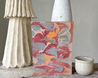 Può includere: Un libro decorativo con copertina marmorizzata con motivi rossi, arancioni, blu e verdi. È appoggiato su una superficie di legno, accanto a un piccolo vaso di ceramica bianco sporco e due oggetti decorativi scolpiti.