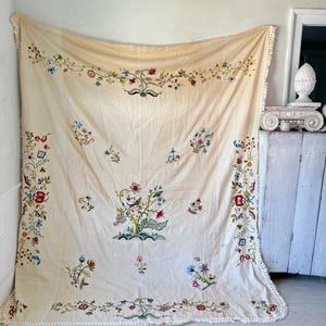 May include: A large, off-white linen tablecloth with a delicate floral embroidery design. The tablecloth features a central motif of a large floral arrangement with a bird, surrounded by smaller floral motifs. The tablecloth has a crocheted border.