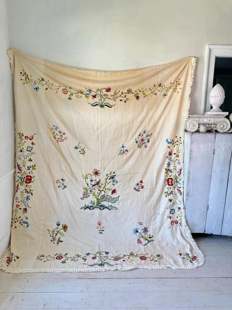 May include: A large, off-white linen tablecloth with a delicate floral embroidery design. The tablecloth features a central motif of a large floral arrangement with a bird, surrounded by smaller floral motifs. The tablecloth has a crocheted border.