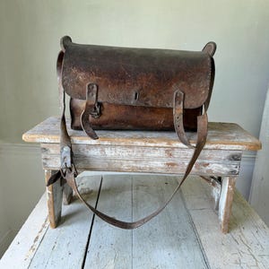 Puede incluir: Un bolso de cuero marrón oscuro con una solapa rectangular, dos correas verticales con hebilla para el cierre y una correa de hombro ajustable. La superficie de cuero muestra una textura desgastada con rasguños y marcas naturales.