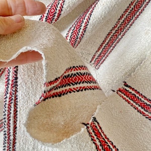 May include: Close-up of a white fabric with red and black stripes. The fabric is woven with a textured pattern and has a frayed edge.