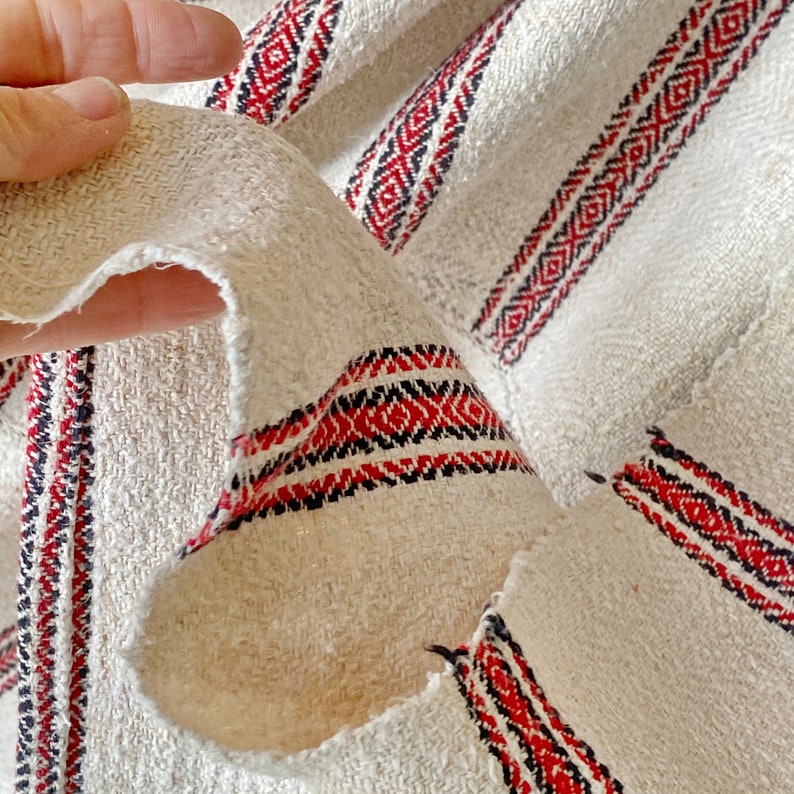 May include: Close-up of a white fabric with red and black stripes. The fabric is woven with a textured pattern and has a frayed edge.