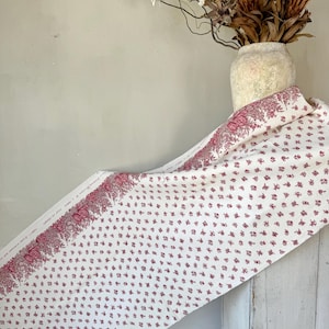 Puede incluir: Tela blanca con un patrón floral rojo. La tela tiene un patrón repetitivo de pequeñas flores rojas y un diseño floral más grande a lo largo del borde. La tela está drapeada sobre una superficie blanca.