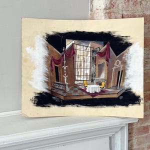 May include: An original watercolor painting of a stage set design. The artwork depicts a detailed interior scene with a table, chairs, and draped curtains. The painting is framed by a black border and has text in the lower left corner.