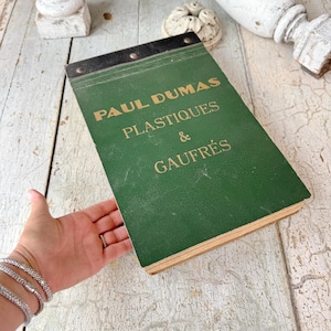 May include: A vintage green notepad with the text "PAUL DUMAS PLASTIQUES & GAUFRÉS" in gold lettering. The notepad has a black binding and is held by a hand with silver bracelets. The background is a distressed, light-coloured wooden surface.