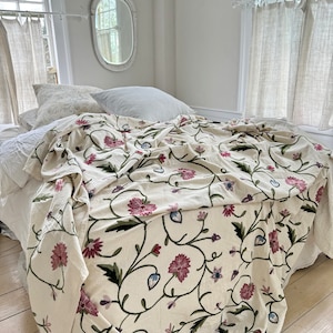 May include: A white throw blanket with an embroidered floral pattern in shades of pink, green, and blue. The blanket is draped over a bed with white linens.
