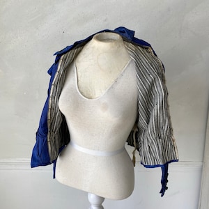 May include: A vintage cobalt blue jacket with a high collar and decorative buttons. The jacket's interior features a gray and white striped lining. The garment is displayed on a white mannequin, showcasing its tailored design and historical style.